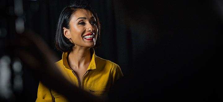 Anita Rani in front of a TV camera smiling off-centre