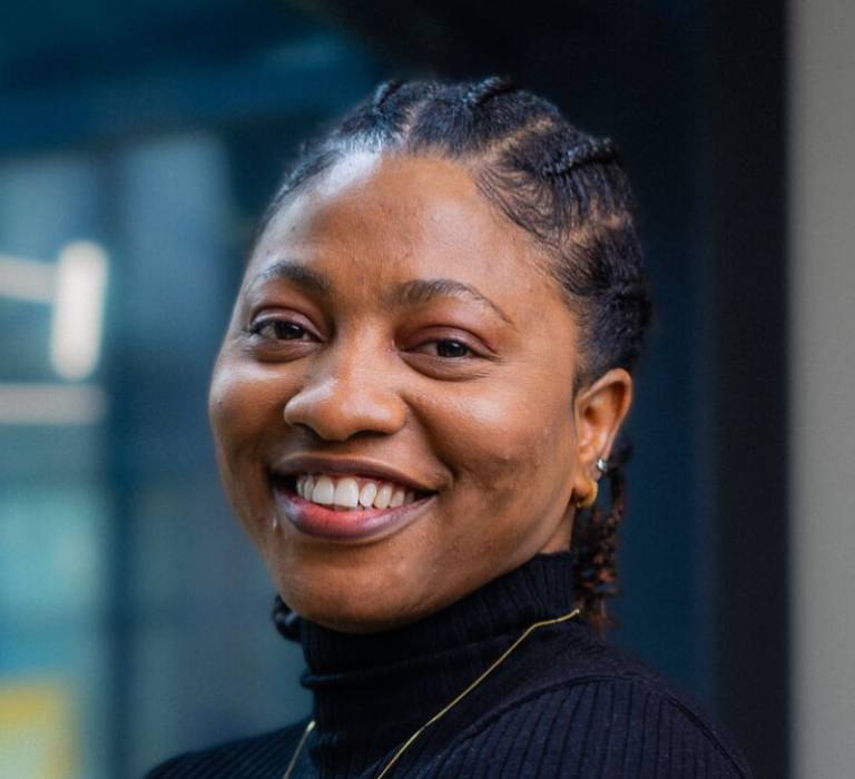 A smiling woman with braids and a roll-neck jumper looks into the camera.