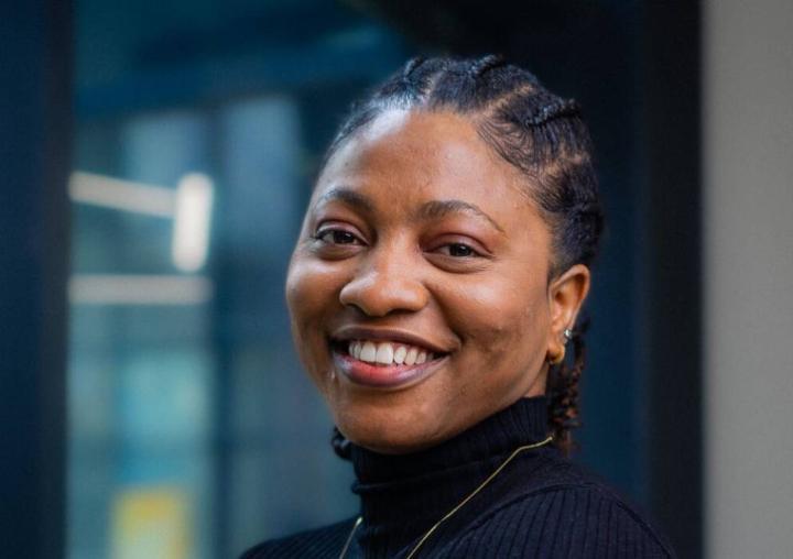 A smiling woman with braids and a roll-neck jumper looks into the camera.