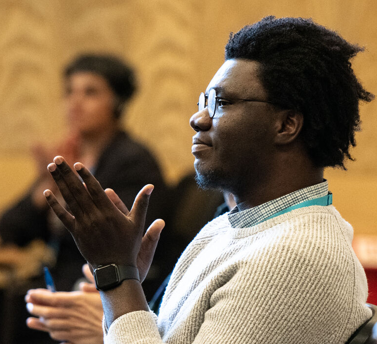 A man with black hair, glasses and a white jumper claps while looking to the left.