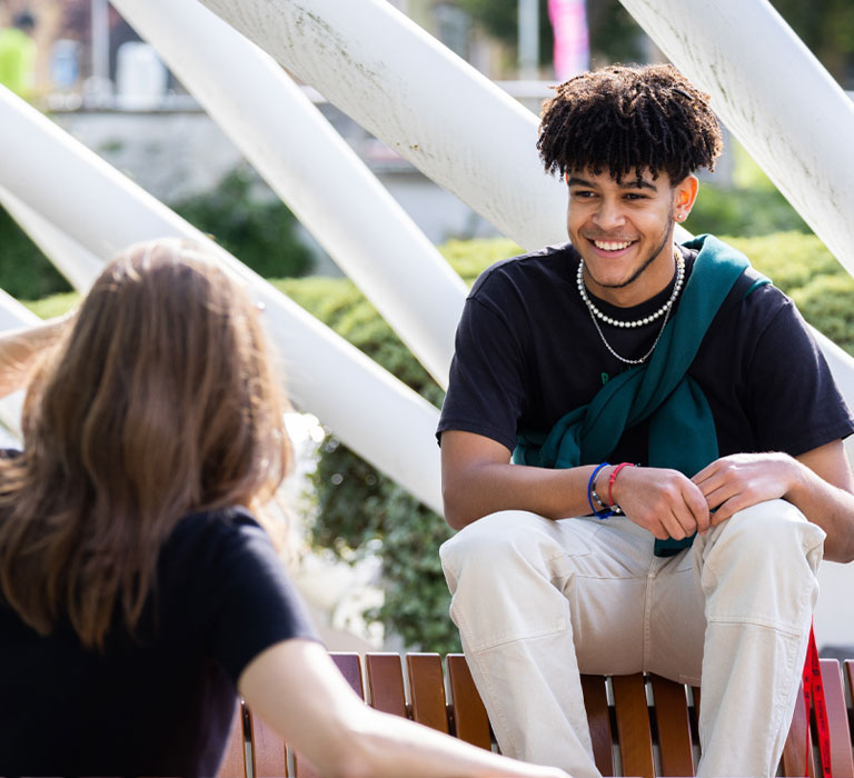 Two students talking to each other, one with back to camera, one smiling at the other outside on campus