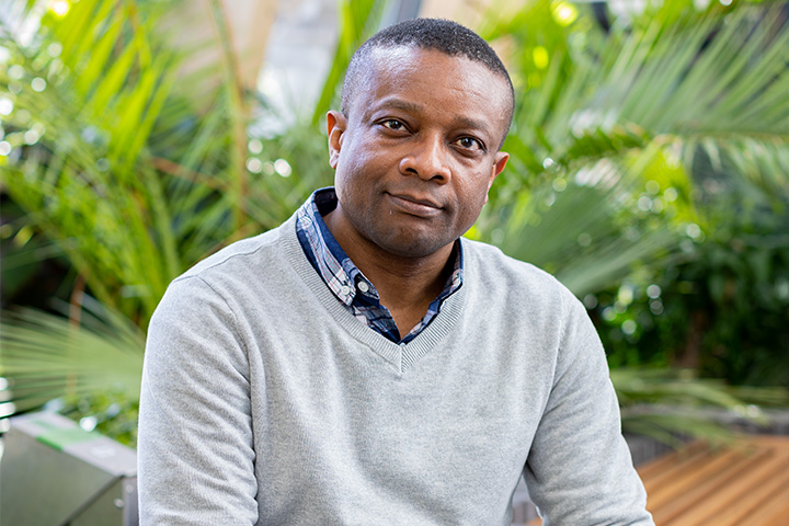 A student wearing a grey sweater sitting on an indoor bench in front of indoor plants resembling palm leaves, smiles at the camera