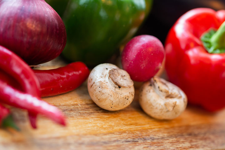 A wooden cutting board with mushrooms, radishes, red onions, red, green peppers and chili peppers