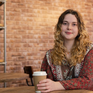 A student smiling at the camera while sitting and holding a takeaway coffee cup