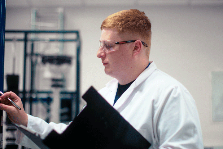 A student wearing a lab coat, safety glasses and carrying a clipboard