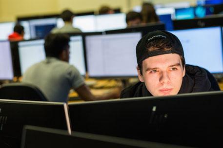 A student sitting in front of two computer monitors, working. More computers with people working at them are in the background.