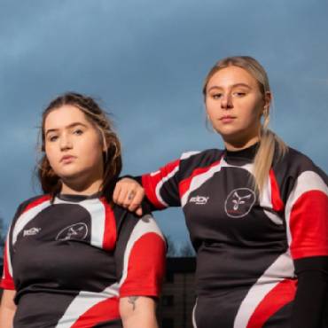 Two women in rugby uniform stand on grass with tough expressions.