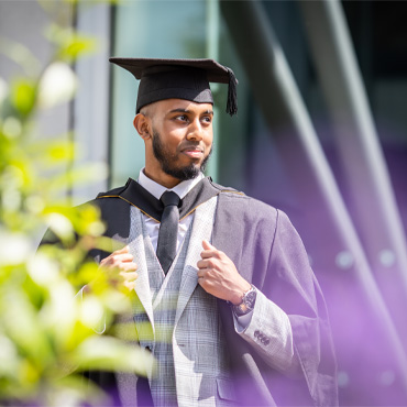 A student posing outside of Richmond Building in a suit and graduation gown and cap.