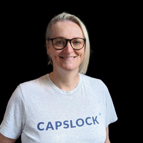 Andrea Cullen, BSc Computing with Management (2000) and PhD Operations and Supply Chain Management (2004) wearing glasses and a grey CAPSLOCK t-shirt, standing against a black background.