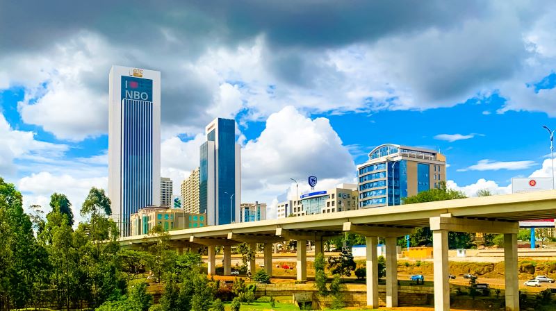 Nairobi skyline which includes tall buildings, under a cloudy sky, with an overpass in the foreground and greenery below.