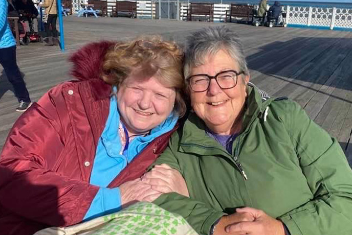 Two people sat down facing the camera, enjoying a cup of tea on the promenade during a trip to the seaside