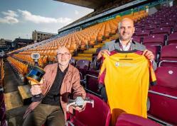 Two people sat in a football stadium. One is holding up a football shirt.