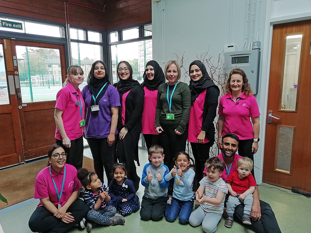 University nursery staff standing behind young children sitting on the floor..