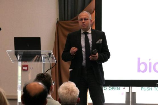 A person holding a mobile phone stands talking on a stage at a conference