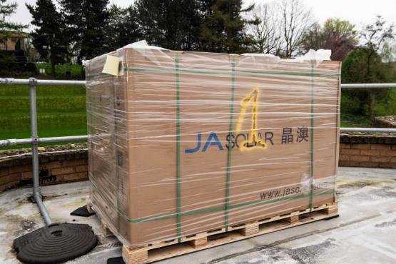 A large wooden pallet covered in see-through film wrap which is filled with solar panels