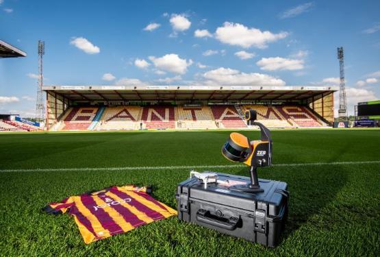 A football shirt lies on the grass touchline of a football pitch next to a box with a drone on top of it inside an empty football stadium