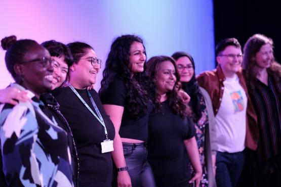 A group of six people stand up next to each other on a stage at a show and all smile