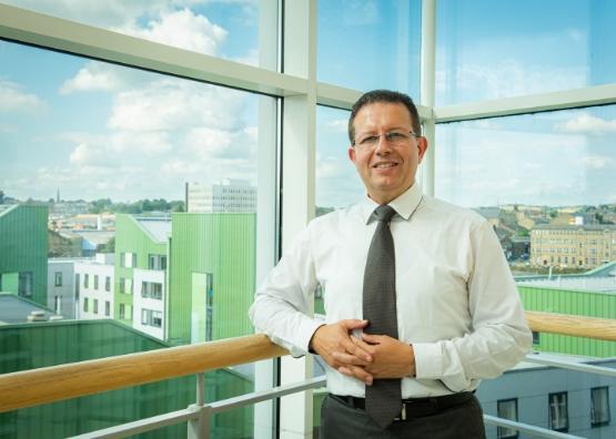 A university staff member wearing a shirt and tie is stood up in front of a glass window in a building