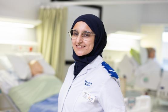 A university student wearing a student nurse uniform in a mock hospital ward with a bed in the background