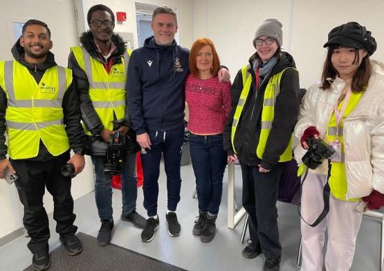 A group of students and staff stand up for photo with football manager wearing a tracksuit