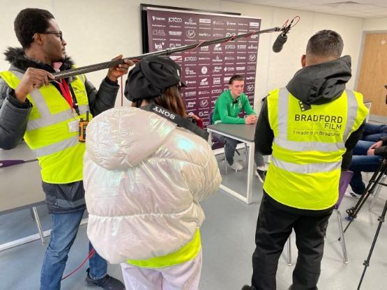 A group of students film a press conference with a football player sat down at a desk