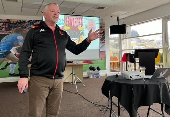 Person in tracksuit top stands up to give guest lecture at sports ground