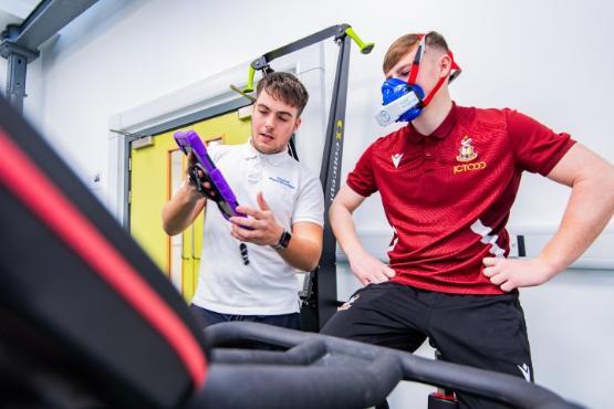 A football player wearing a tracksuit on an exercise bike while wearing a face mask gets a demonstration from a student holding a tablet computer