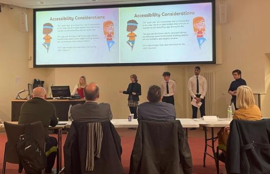 A group of students in the background stood up delivering a pitch to people sat at a table in the picture's foreground