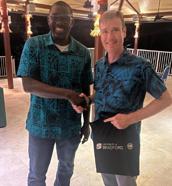 Two people stand next to each other and shake hands while one holds a University of Bradford tote bag