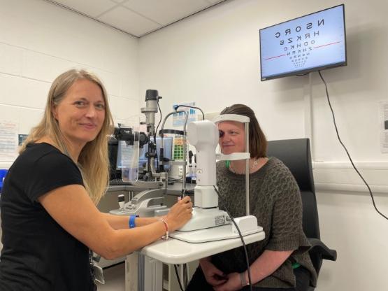 A person on the left in charge of a mock eye test as another person leans forward on testing equipment