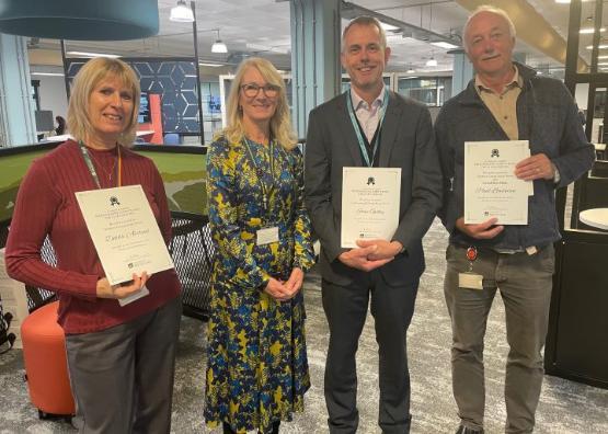 Four staff members stand next to each other three of them holding certificates in a new library area