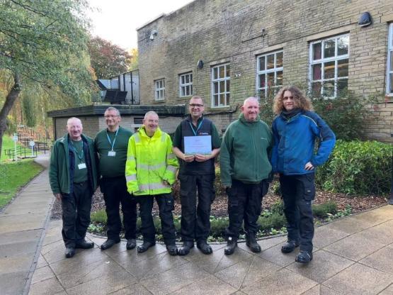 A group of six university gardeners stand up next to each other in front of building in an area of green space