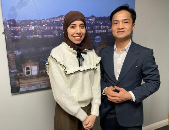 Two students stand next to each other with a photograph in a frame behind them