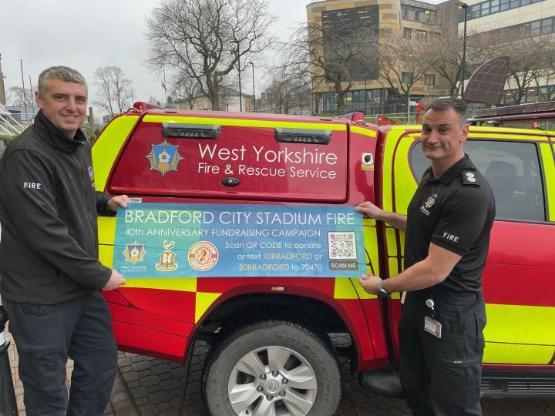 Two firefighters stand at opposite ends of a marked fire vehicle holding a banner for a charity appeal