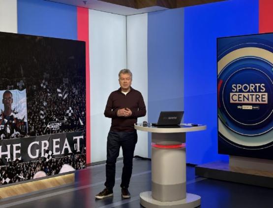 A presenter stood up with a small podium to their left in a studio with big screens either side of them