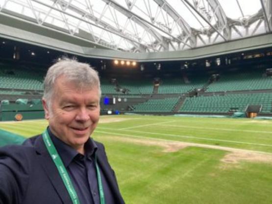 A person in the foreground takes a selfie photo in a grass court tennis stadium