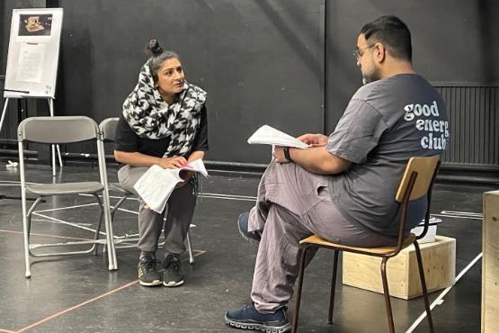 A play rehearsal photo of a woman wearing a headress pleading with a man also sat down