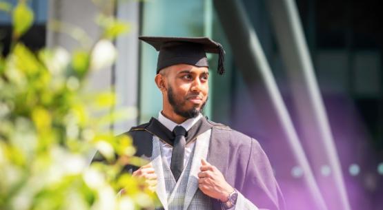 A male graduate wearing a mortarboard and gown looking pleased