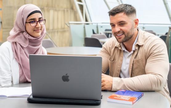 Two former students sit next to each other at a table looking at a laptop on a table in front of them