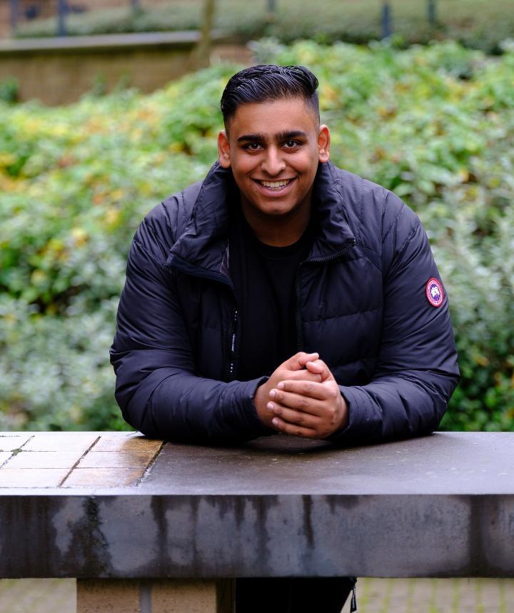 Portrait shot of Zubayr sat at a stone bench in front of some greenery.