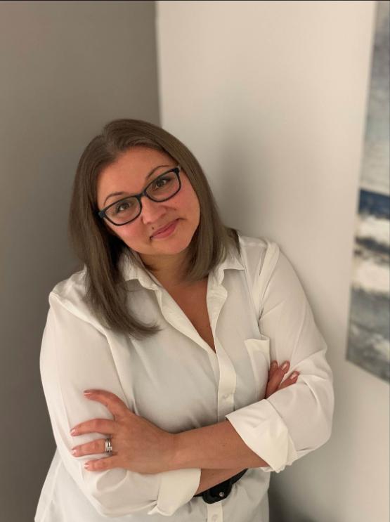 Tatjana wearing stood against the corner of a room with her arms folded and head leaning to one side.