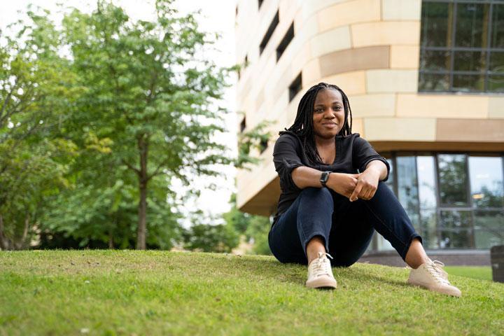 A person sitting on a small mound of grass, smiling at the camera, with a tree and building in the background.