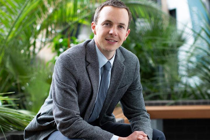 A person sitting on a bench and smiling at the camera with plants in the background.