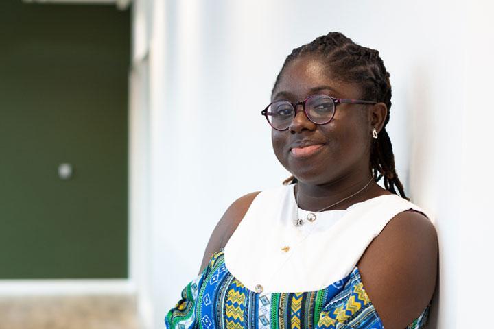 A person in a well-lit indoor space, leaning against a wall and smiling at the camera.