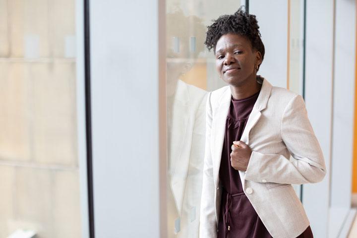 A smartly dressed person in a white jacket leaning on a window and smiling at the camera.