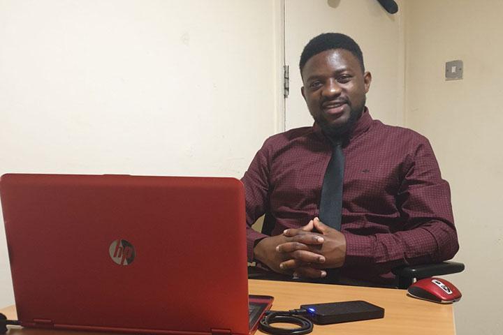 A person sitting at a desk, dressed smartly and smiling at the camera. There is a laptop on the desk.