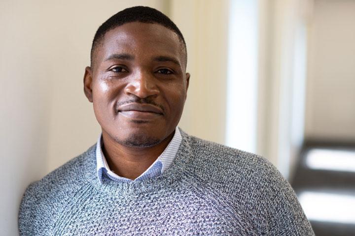 A smartly dressed person smiling at the camera and leaning casually against a wall.