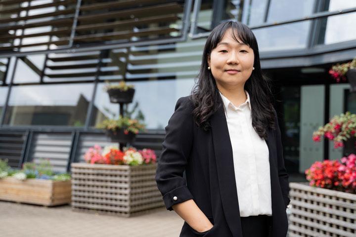 A smartly dressed person standing outside and smiling at the camera. There is a glass-fronted building in the background.