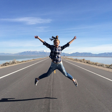 A student jumping joyfully in the middle of an empty road, surrounded by lakes and mountains.