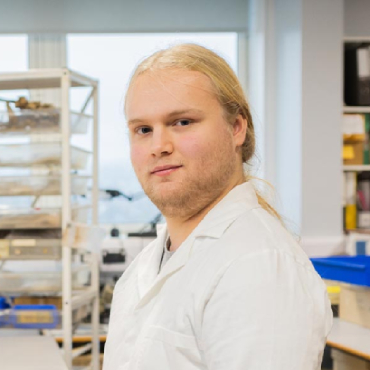 A student with long, fair hair and a white coat looks into the camera with archaeological equipment behind them on white shelves.